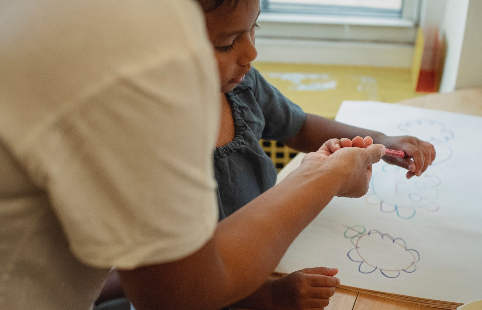An adult passing a crayon to a child drawing at a table by a window